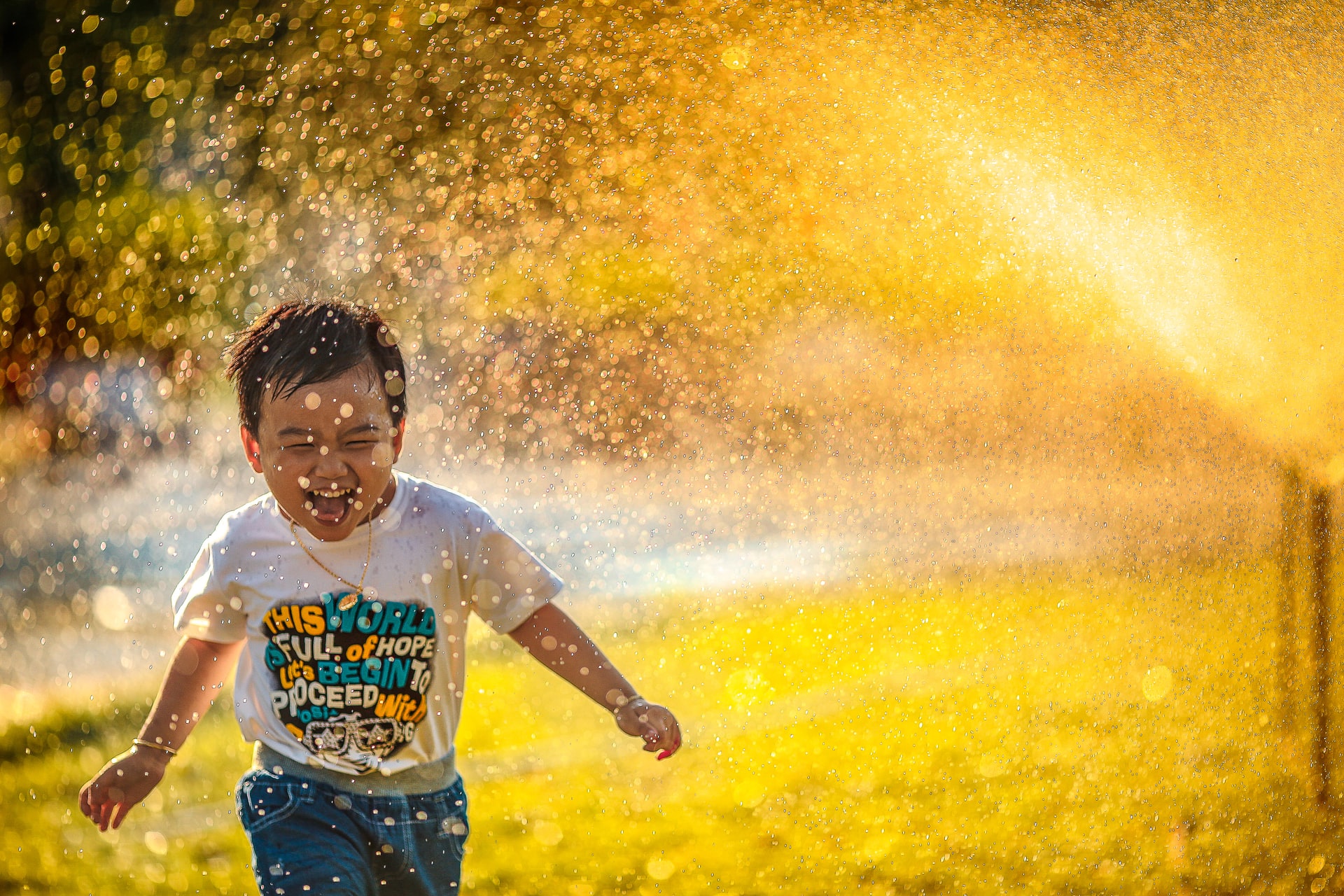 A small boy is getting drenched by the water splashin a garden and he is so happy about it
