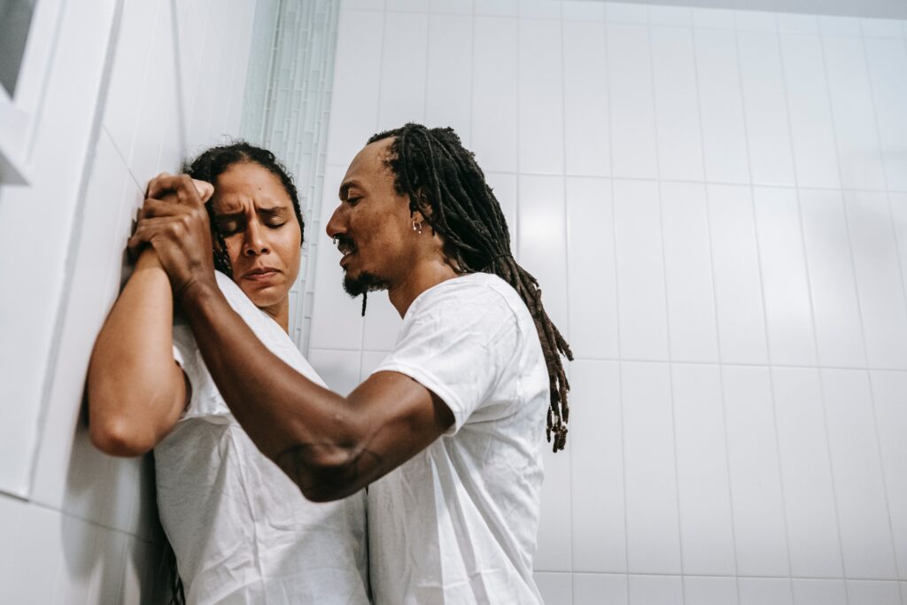 A man has hela a woman to the wall and is hurting her by clasping her hands tightly. She looks in pain.