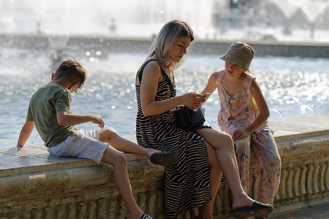 A Mother looking at her phone sitting on a lakefront with her two children, a boy & a girl